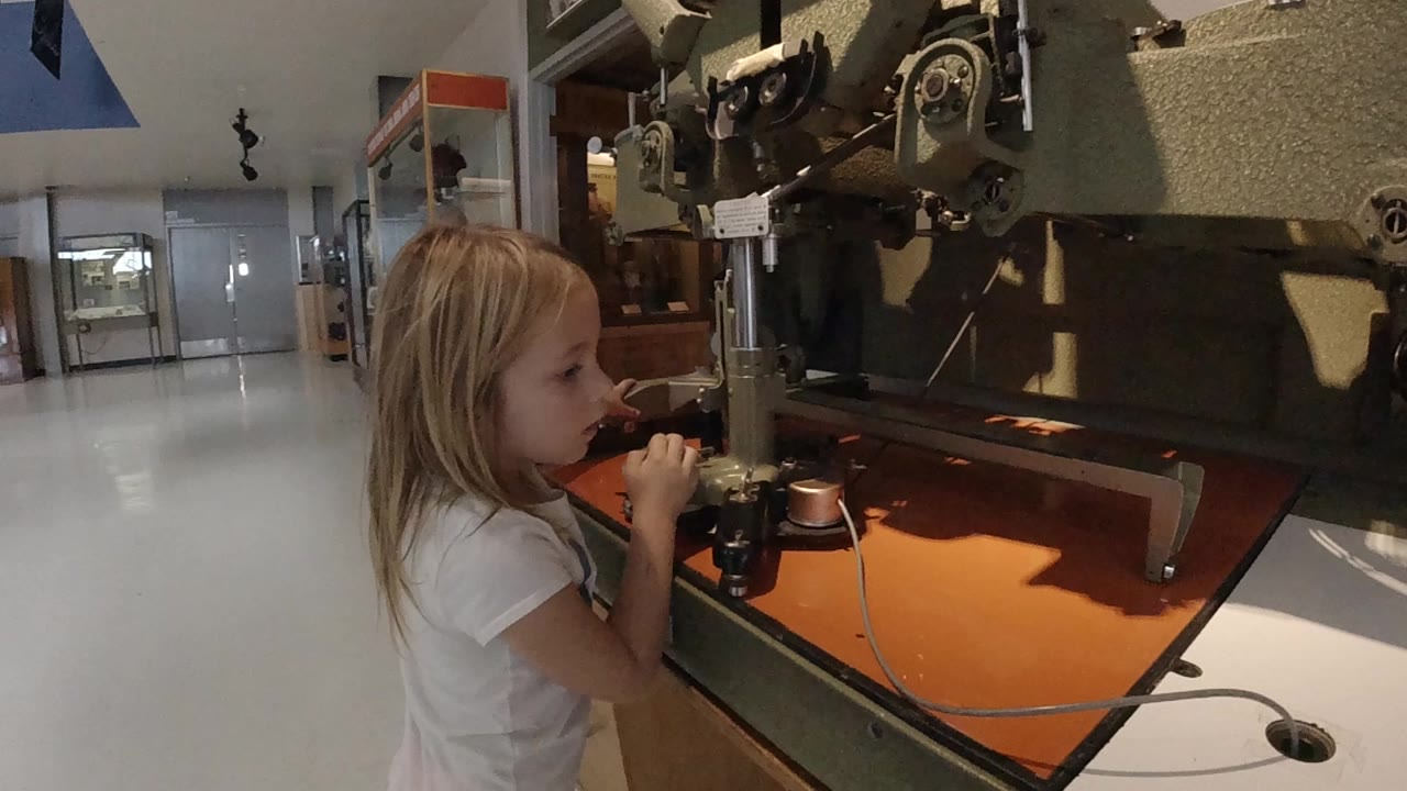 At the start of the Colorado clip, a blonde child stands close to a large mechanical display with an orange tabletop surface and a bright hallway behind her.