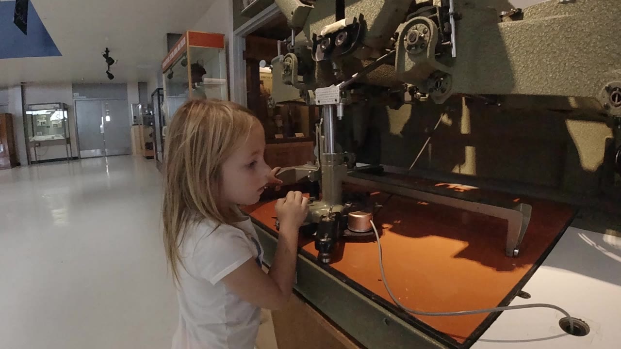 The girl leans toward the black controls on the large machine while the orange surface and metal assembly dominate the right side of the Colorado interior frame.