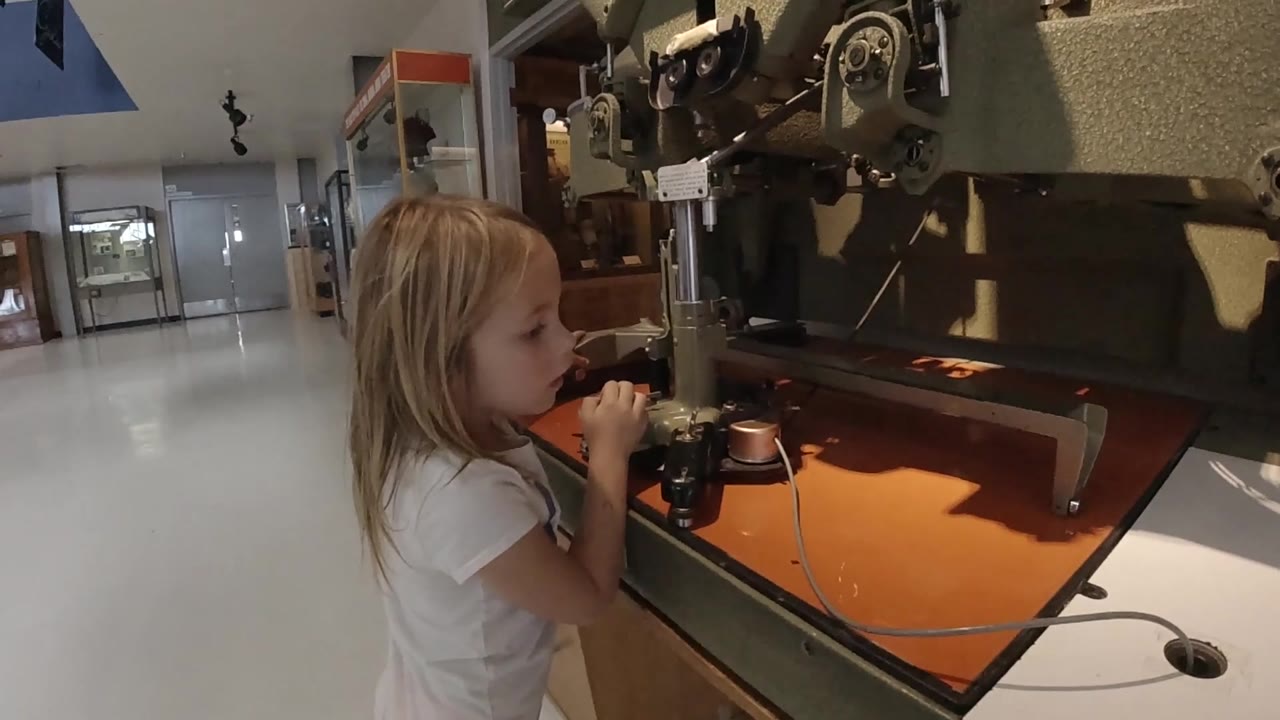 The child remains beside the machine, with her face in profile, hands near the controls, and the orange work surface catching warm light in the foreground.