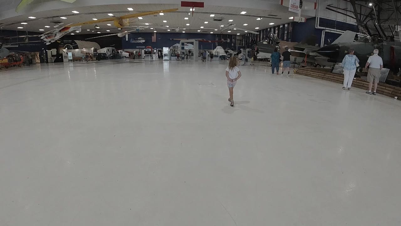 A little girl stands near the center of a broad pale floor inside a Colorado museum-like hall, with aircraft exhibits and visitors visible in the background.