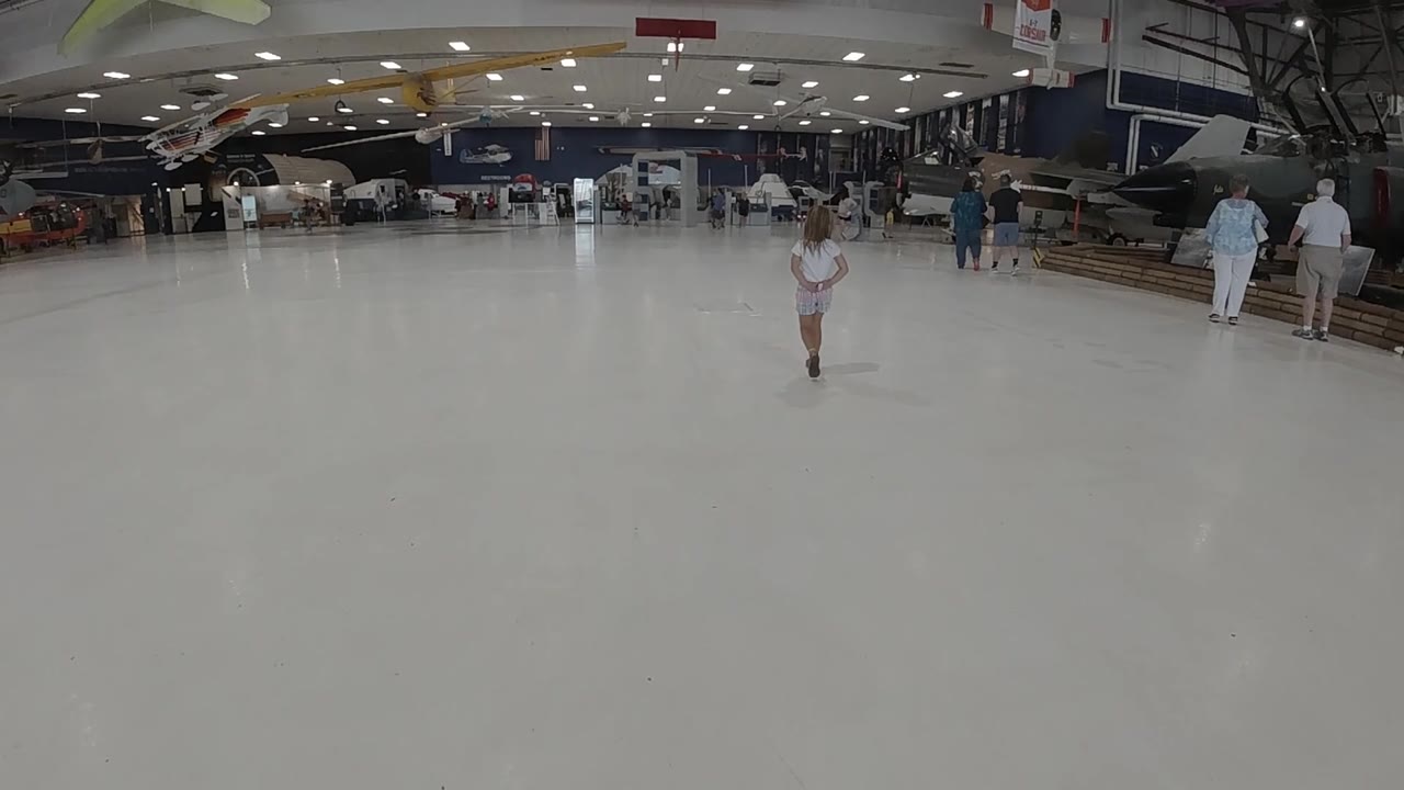 A little girl walks toward the camera across the pale floor, with aircraft shapes, display areas, and several people across the back wall of the Colorado hall.