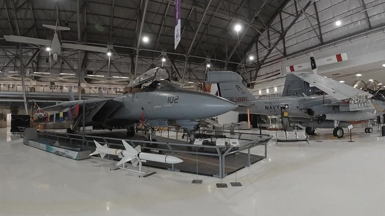 Wide interior of a military aviation museum in Denver showing a large dark jet aircraft on static display, white missiles on low stands in the foreground, steel truss ceiling with bright overhead lights, and no people visible.