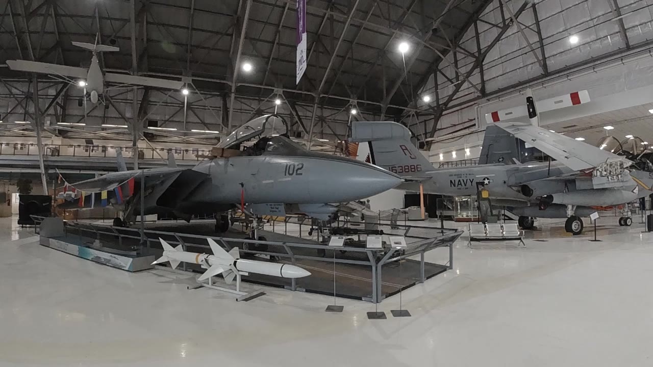 Military aviation museum interior in Denver with a large dark jet on static display center-left, white missiles in the foreground, and a high steel-truss ceiling with overhead lighting; no people visible.