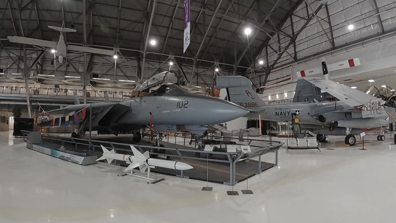 Museum hangar interior showing a large dark jet aircraft and white missiles on static display stands in Denver, steel ceiling trusses and spot lighting overhead, no people visible.