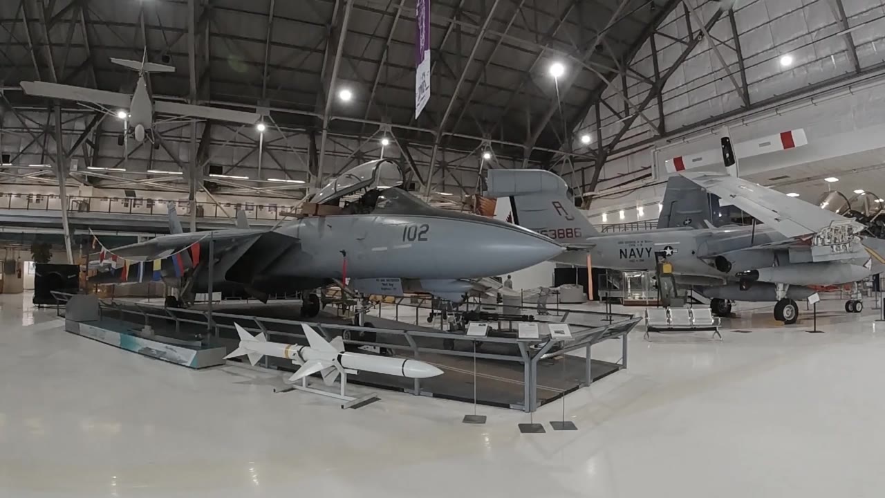 Dark military jet aircraft on museum display with white missiles in foreground and industrial hangar ceiling overhead.