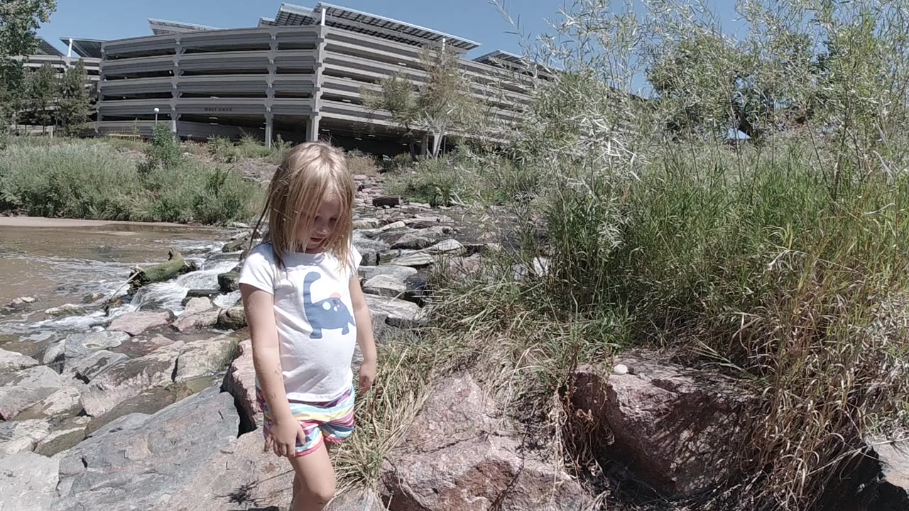 A young blonde girl stands on boulders beside a muddy stream in Denver, with tall grass and a concrete parking structure behind her.