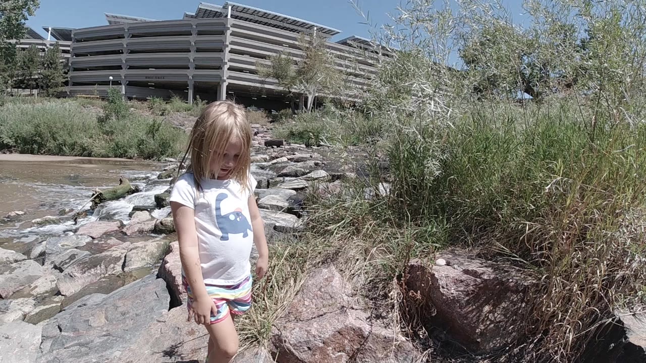 The girl looks down at the uneven rocks while standing near tall streamside grass in Denver, with the parking structure across the water.