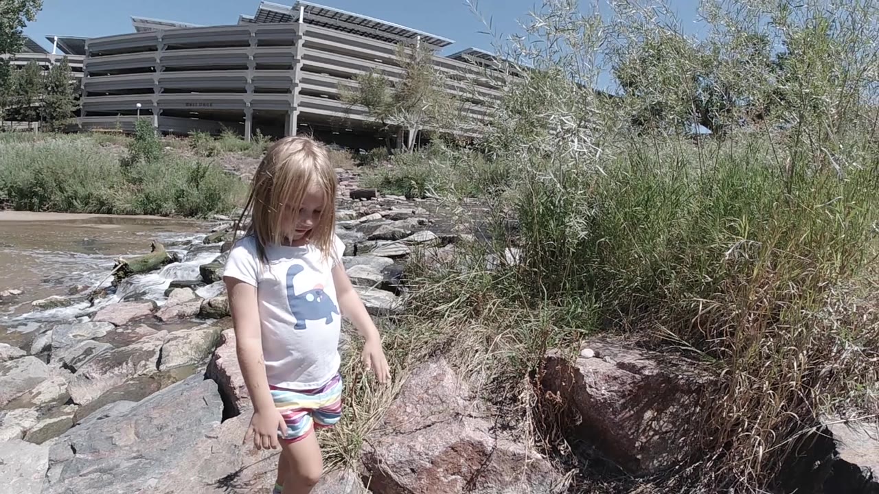 The child shifts her stance near large pale rocks beside the stream, framed by brush and a concrete structure in the Denver background.
