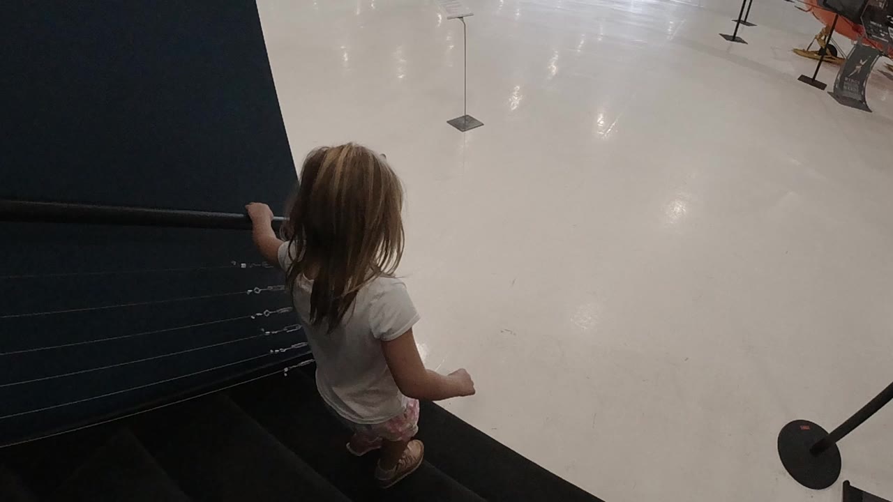 A young blonde girl in a white shirt stands near the top of a dark staircase in an indoor Denver public display space.