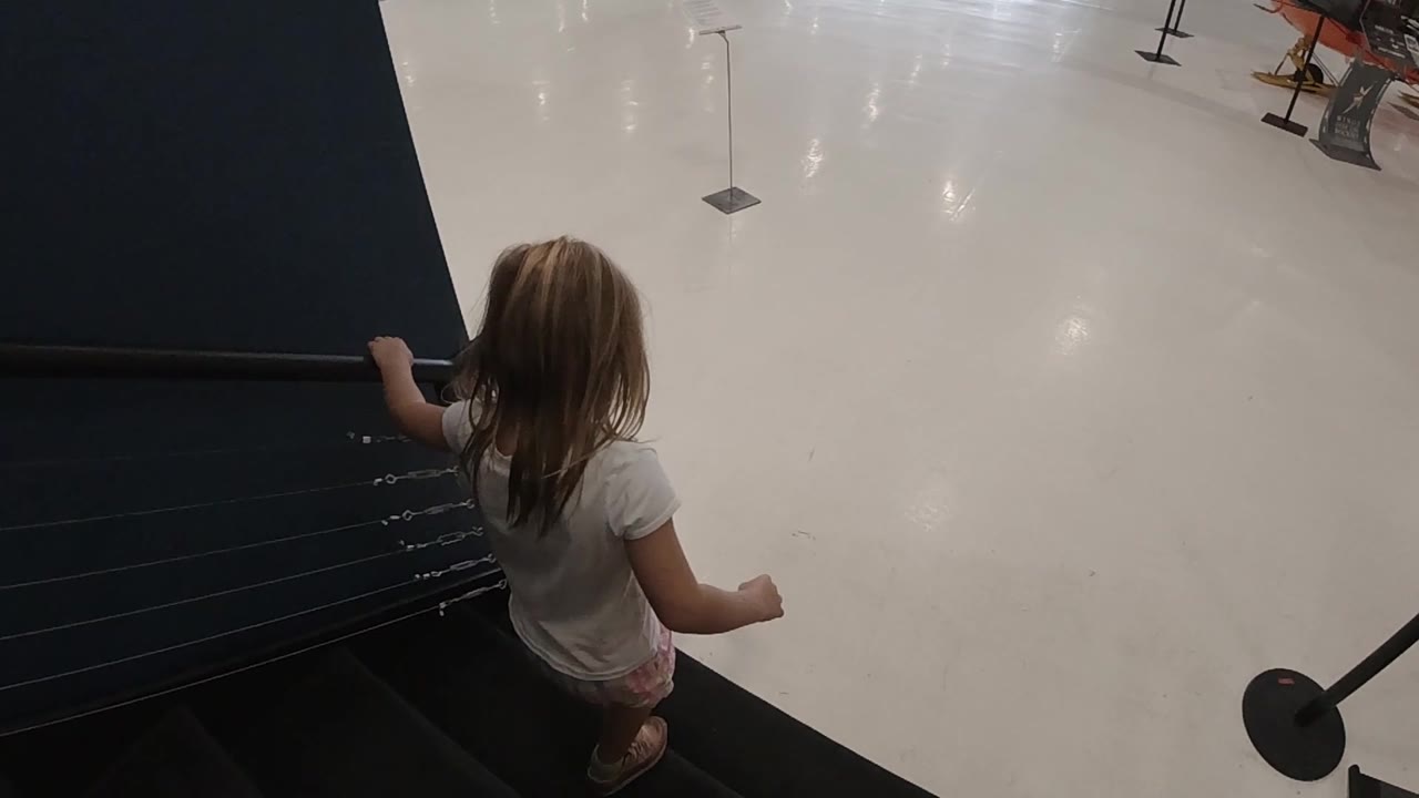 The child continues down beside a dark wall, with the glossy hall floor opening below and display stanchions in the distance.