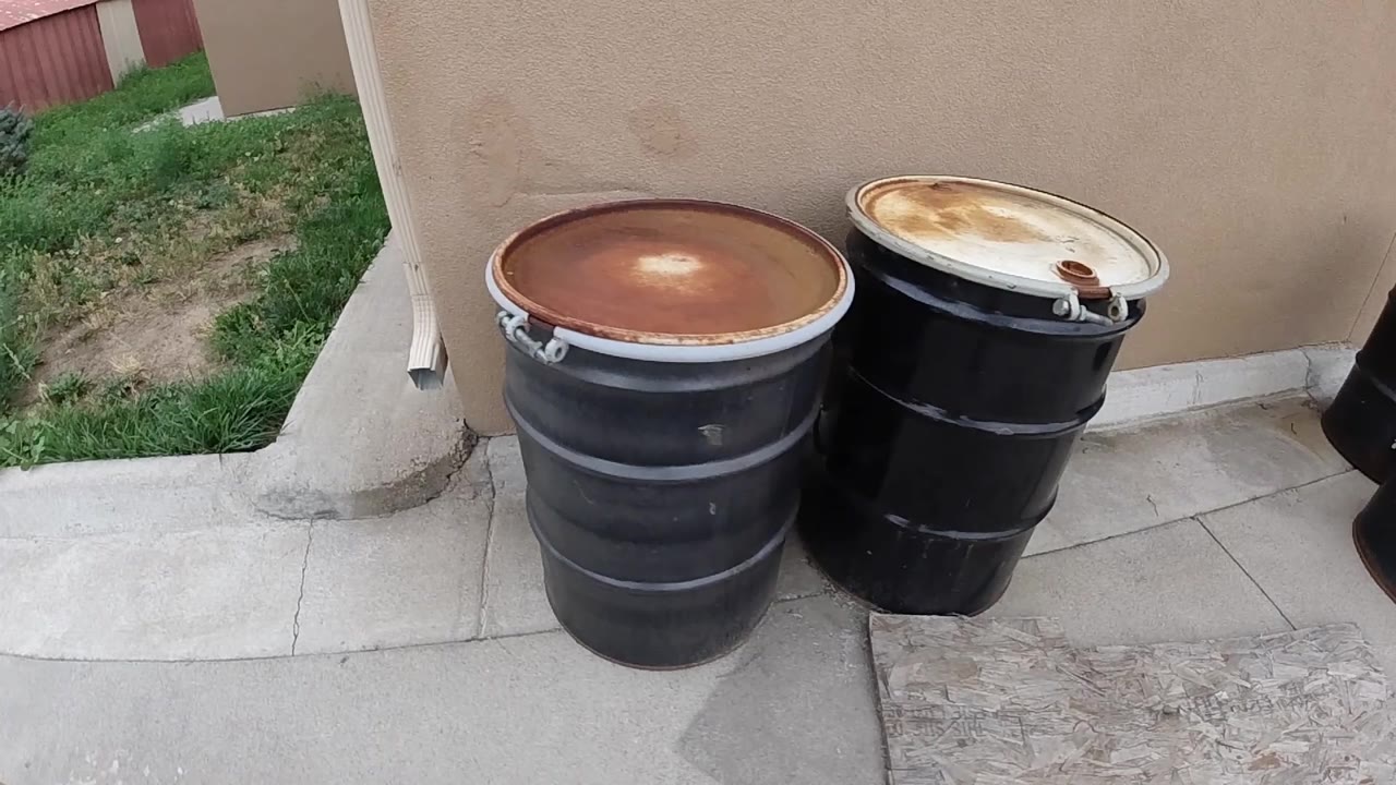 Two black corroded 55-gallon steel drums on a concrete pad against a tan stucco building wall in Denver, Colorado; no visible people