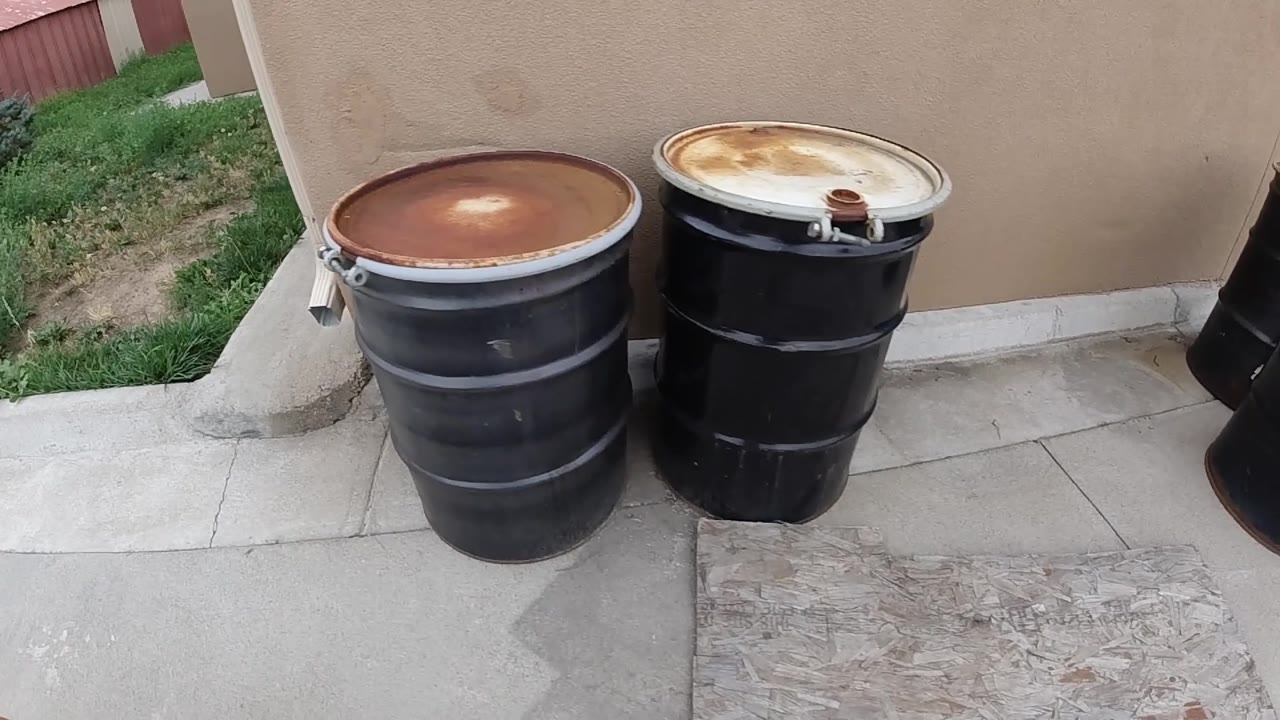 Three black steel 55-gallon drums on concrete, with a third barrel becoming visible on the right side of frame