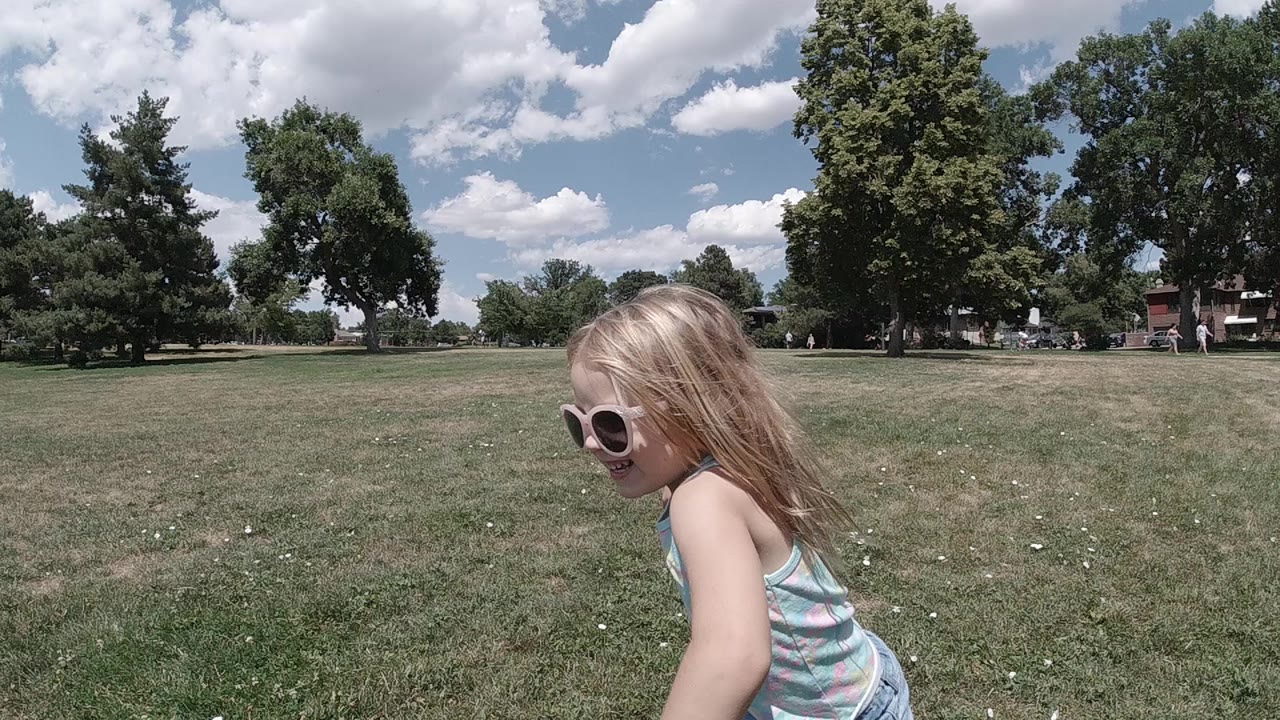 Young blonde girl in a light blue sleeveless top runs left to right across an open flat grass park field; a row of mature deciduous trees lines the horizon under a blue sky with white clouds; no other people are visible in the frame