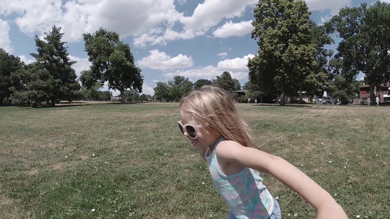 Young blonde girl in profile view runs across an open park grass field, her long blonde hair flying; mature deciduous trees line the background and blue sky with scattered clouds is visible above; no other people in frame