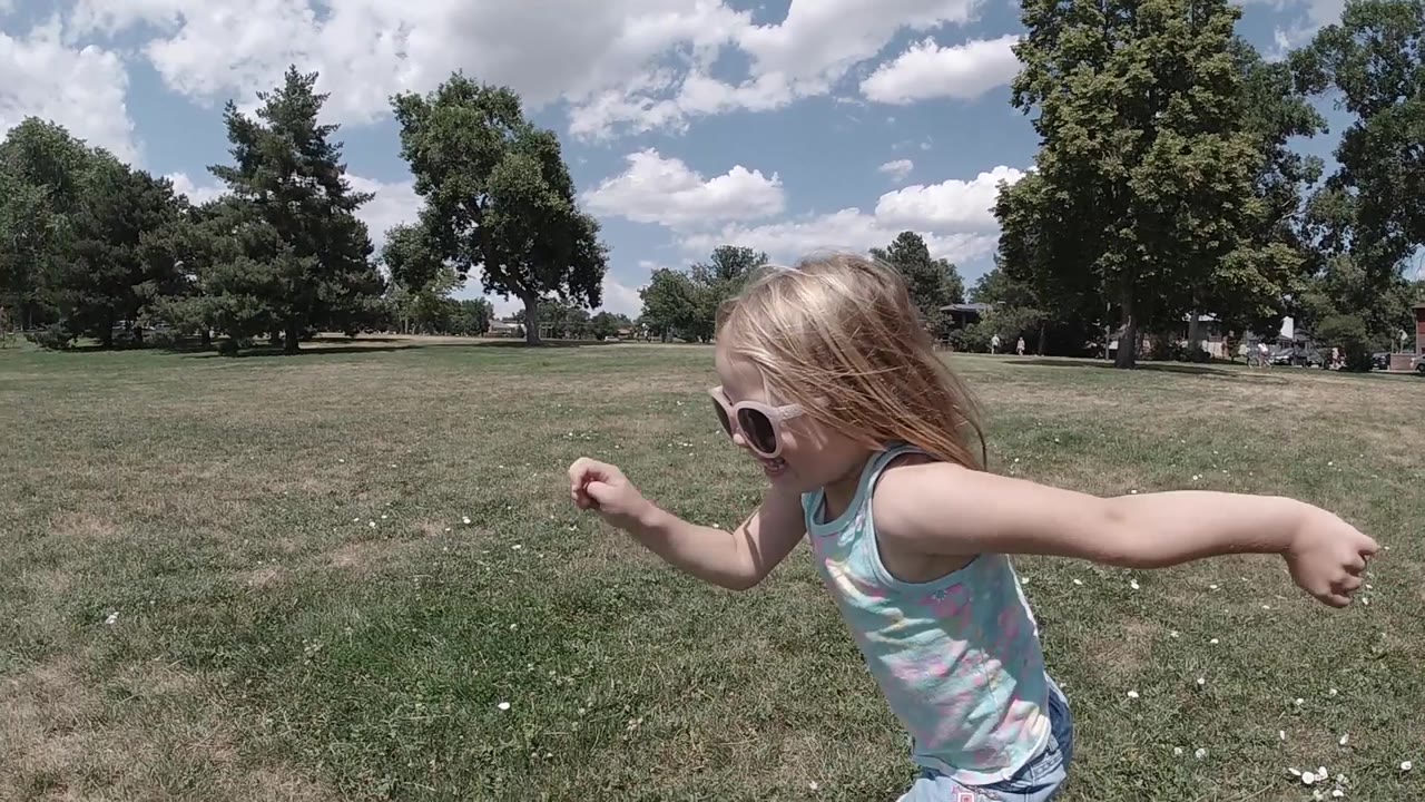Young blonde girl in light blue top running through park, hair extended in motion, trees and sky visible