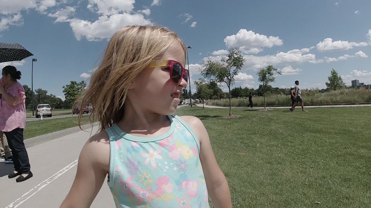 In Denver, a young blonde girl in red sunglasses walks close to camera beside a sidewalk, with grass, trees, clouds, parked cars, and adults behind her.