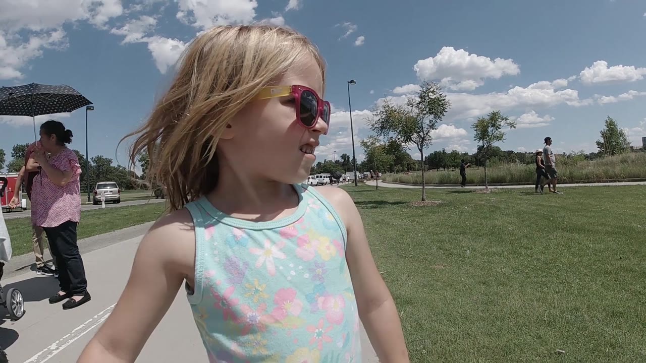 The child walks in the foreground at a Denver park while adults, a stroller, parked cars, grass, small trees, and scattered clouds fill the background.