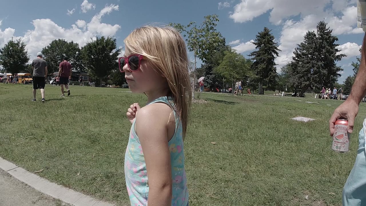 The girl remains in left foreground in Denver, facing sideways as she walks past the lawn while a partial adult arm with a bottle appears at the right edge.