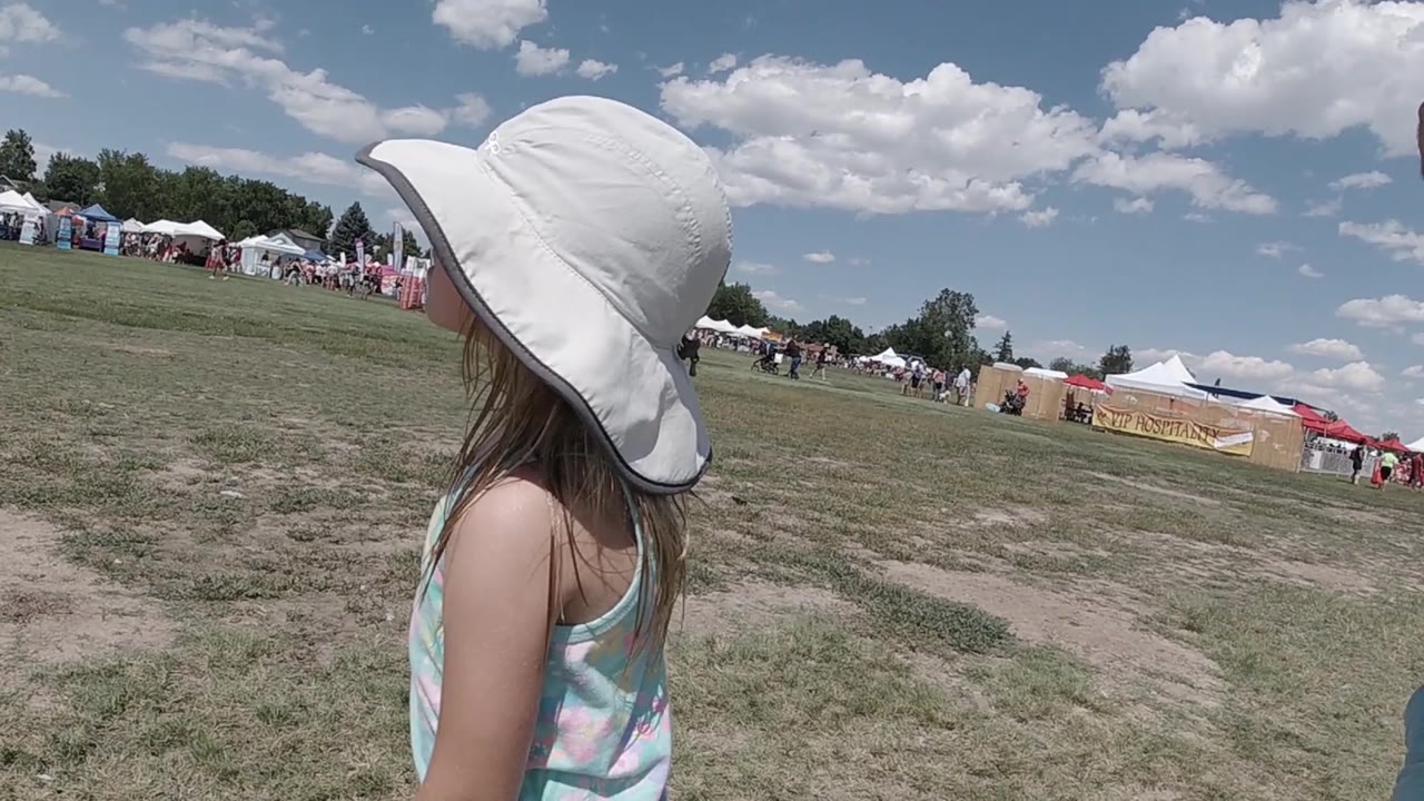The child remains turned away in the Denver festival field, with vendor tents and small groups of people lining the far side of the grass.