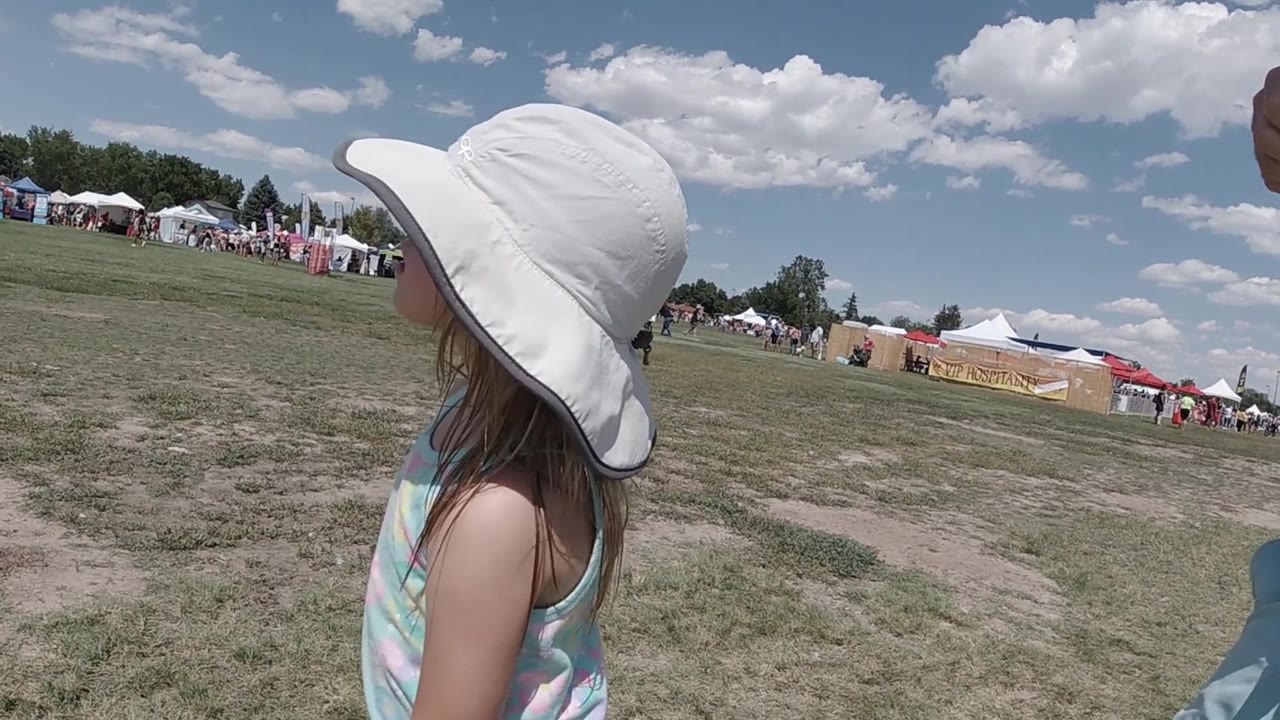 The girl continues across the foreground in Denver, her white hat filling the upper frame while tents, people, and a bright clouded sky sit behind her.