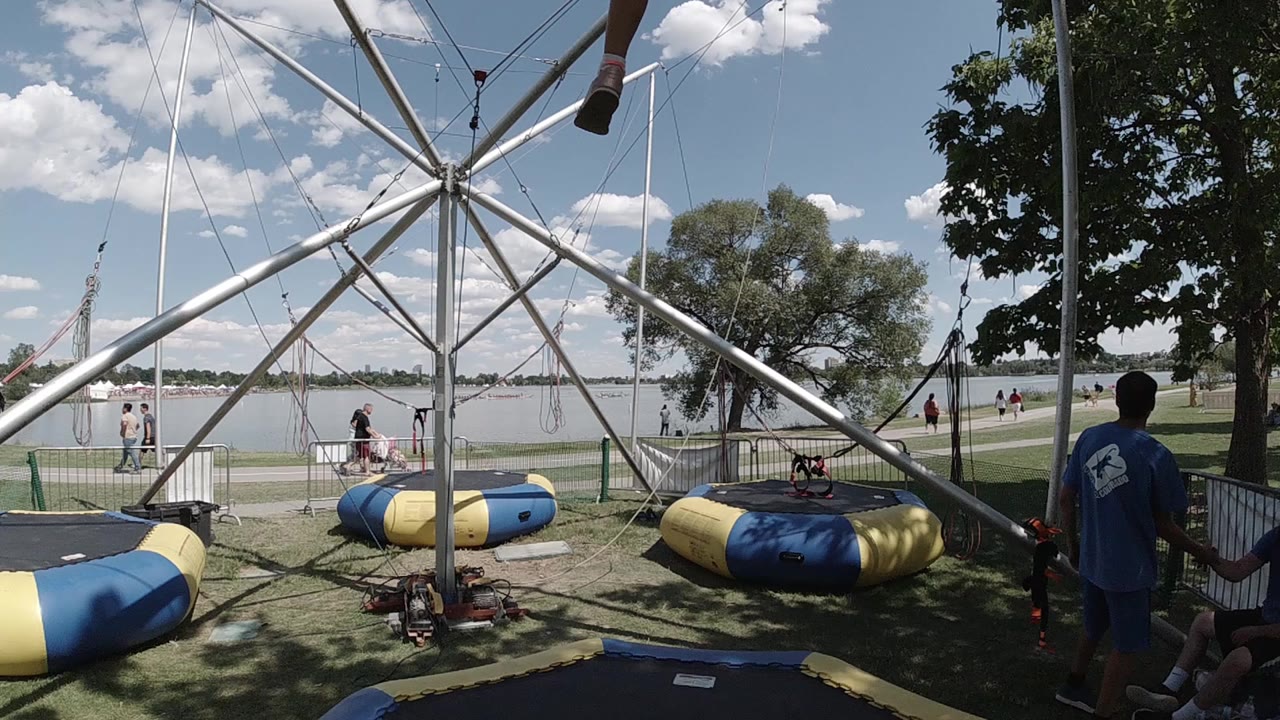 Wide Denver park view of a bungee trampoline setup before the rider enters, with blue and yellow pads, metal supports, trees, water, and an attendant at right.