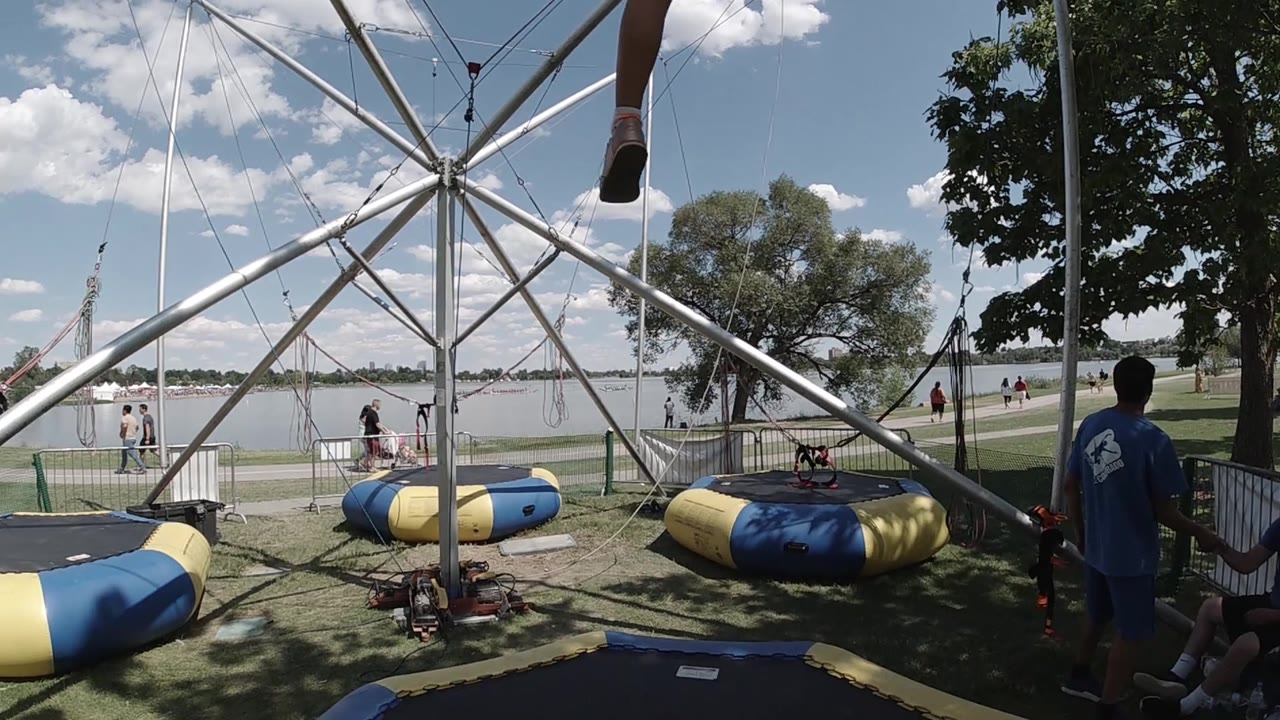 A wide view holds on the bungee trampoline structure in Denver, with empty landing pads, open grass, water behind a fence, and the attendant at far right.