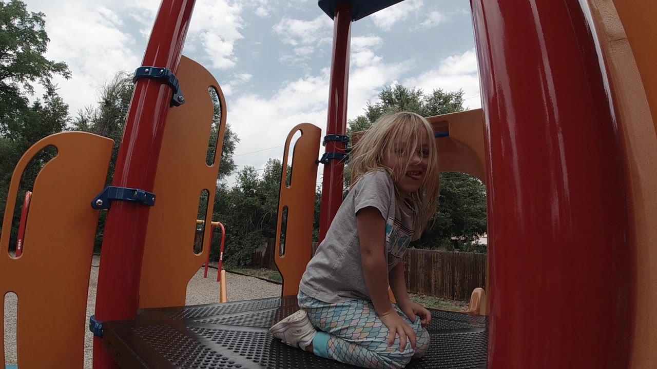 The child leans forward on the black perforated platform in Denver, framed by red vertical supports and orange playground panels.