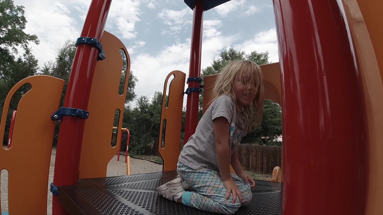 A young girl kneels near the right side of the playground platform, with red posts, orange panels, trees, fence, and cloudy sky visible in Denver.
