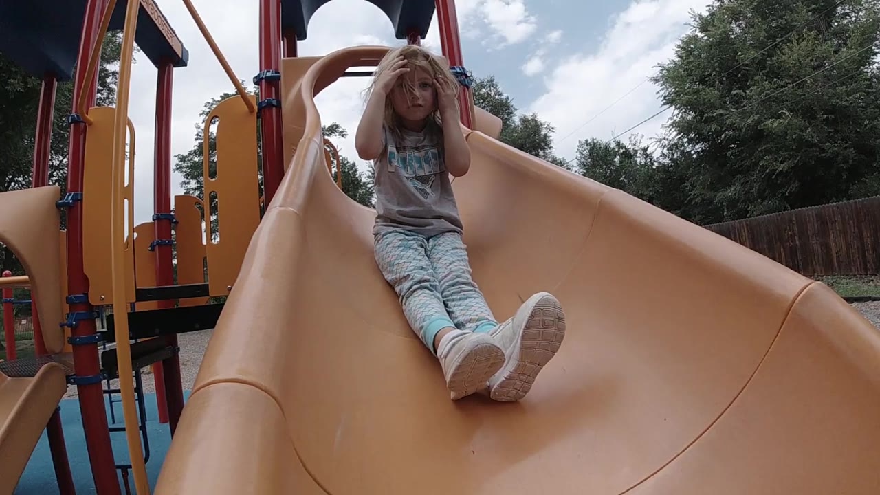 A young blonde girl sits near the upper section of a tan playground slide in Denver, with one hand near her hair and trees behind the play structure.
