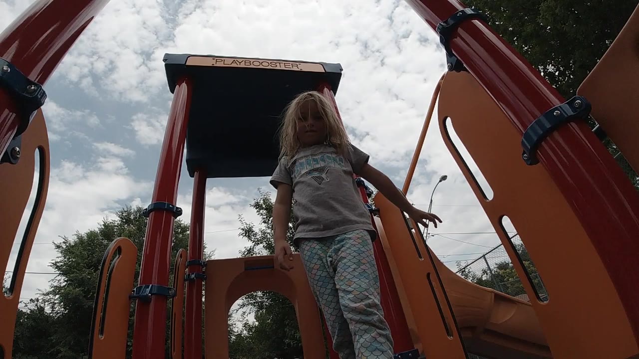 A blonde young girl stands on orange playground equipment in Denver, framed from a low angle under a bright clouded sky.