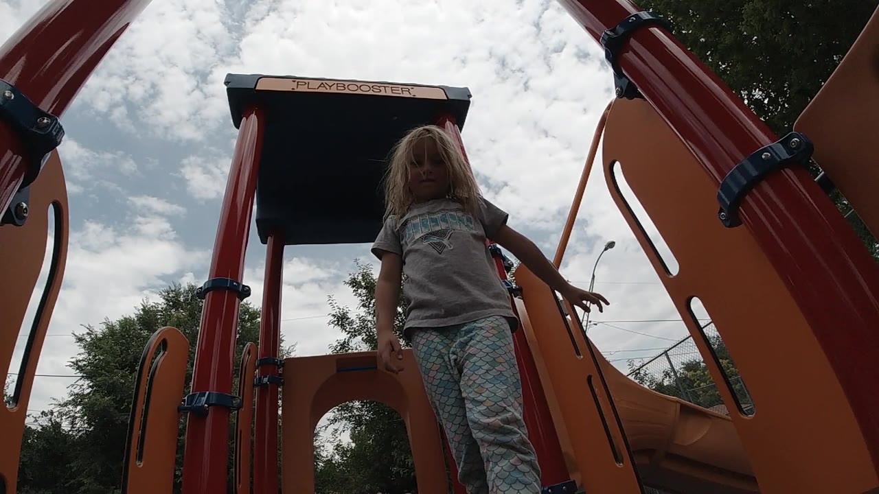 The girl remains centered between red playground posts, with her arms near the rails and clouds filling the sky behind her.