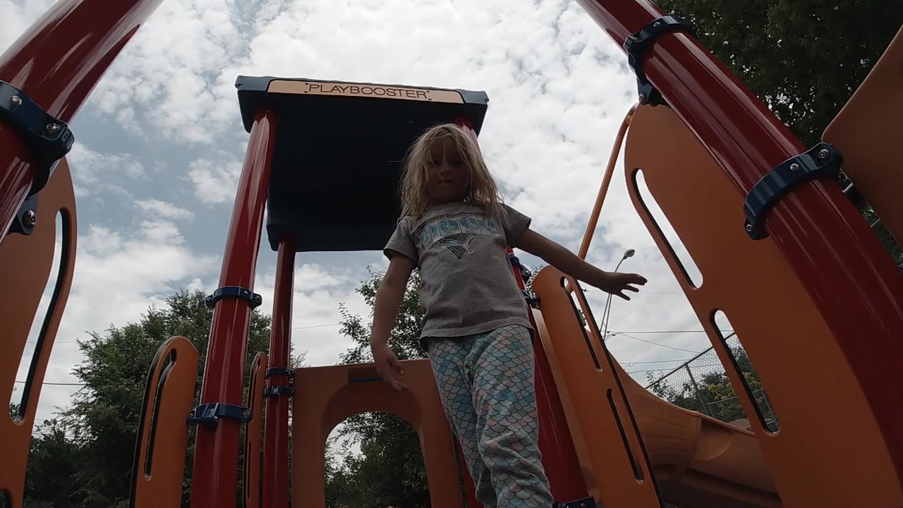 A young blonde child stands on the elevated playground platform, bordered by orange side panels and red posts.