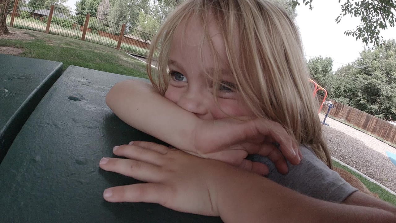 At the start of the Denver clip, a young blonde girl rests her chin and forearms on a dark green picnic table with grass, fence, and trees behind her.