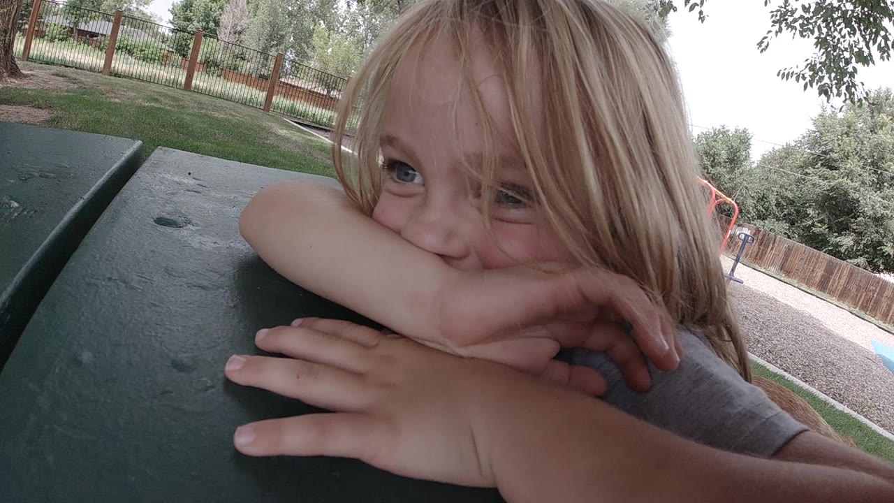 The girl looks upward from behind her forearm while leaning on the green picnic table, with the park fence and trees soft in the background.