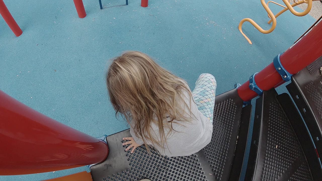 Overhead view in Denver of a young blonde child sitting beside a red playground slide on black perforated decking with blue rubber ground below.