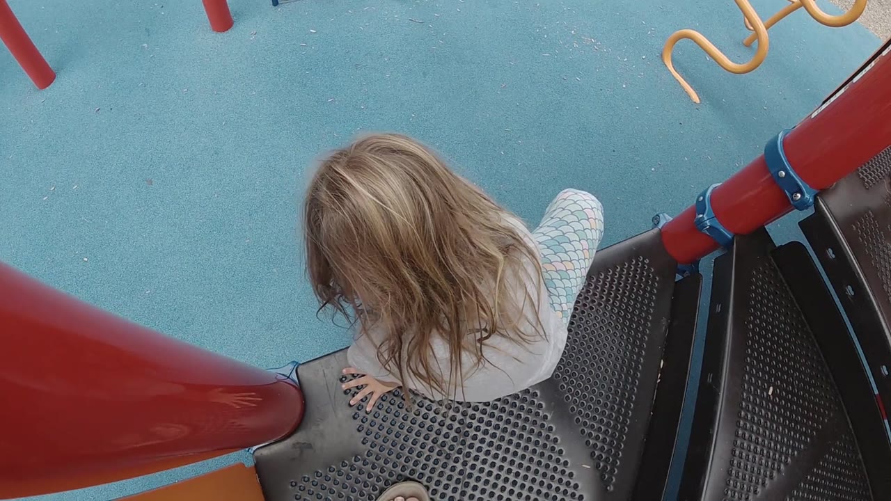 The child sits at the edge of the black playground platform in Denver, with one hand near the deck and the red slide tube cutting across the left side of frame.