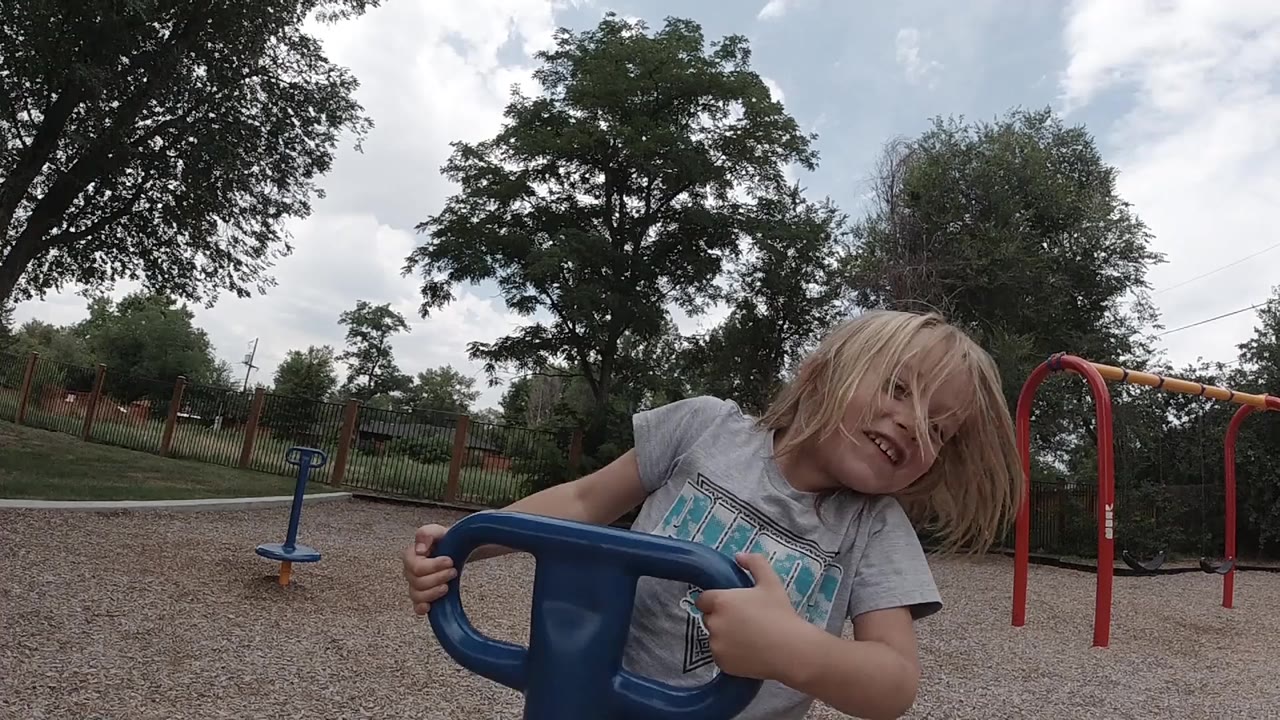 A blonde young girl in Denver grips a blue spinning playground handle and leans toward the camera, with trees, gravel, and red playground bars behind her.