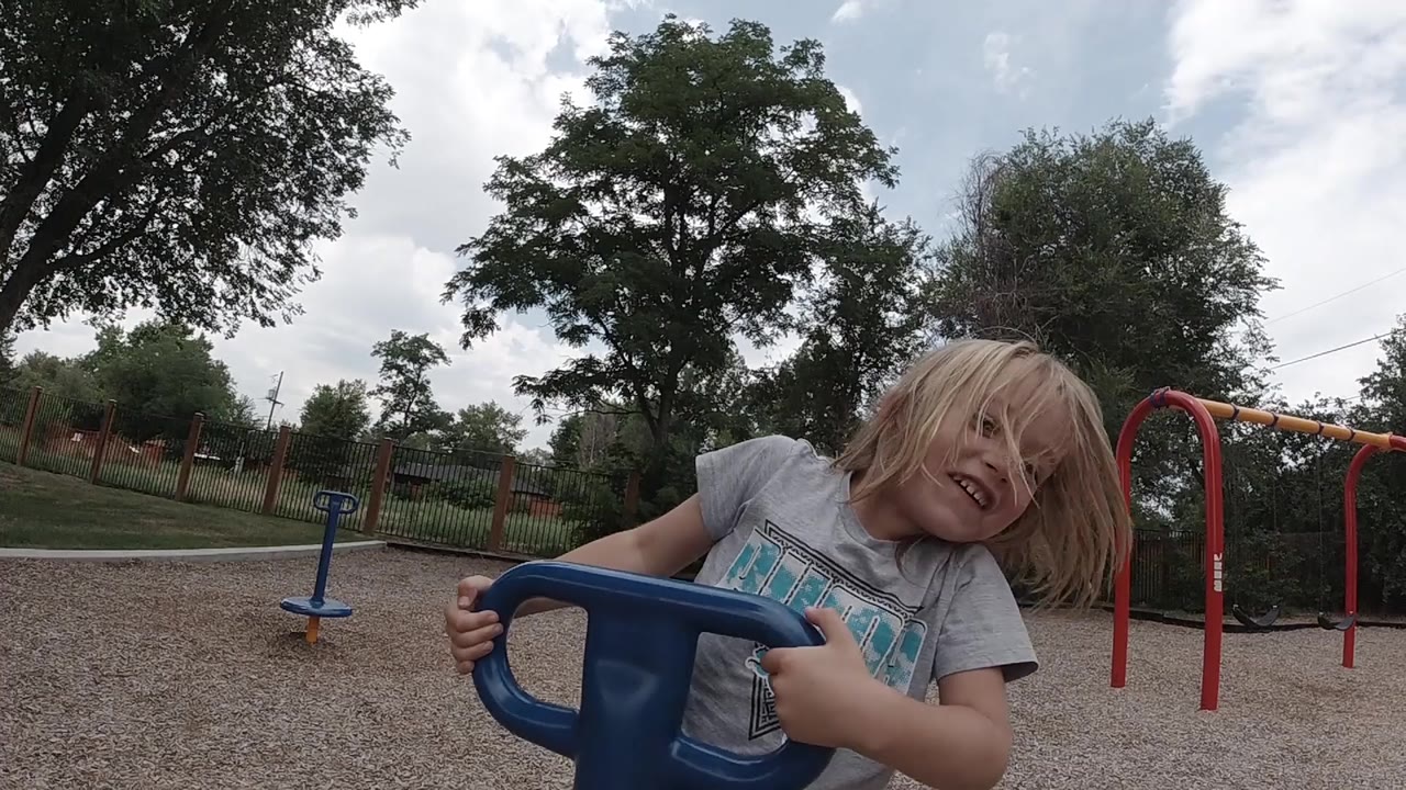 The young girl smiles while holding the blue spinner in a Denver park, with her hair partly over her face and red play equipment in the background.