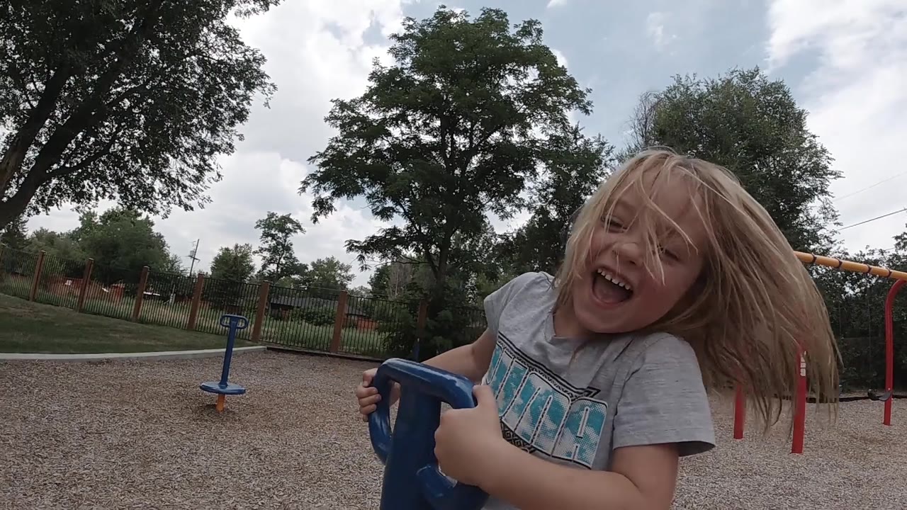 A laughing blonde child holds the blue playground spinner near the camera as wind lifts her hair, with gravel and red bars visible in the Denver park.