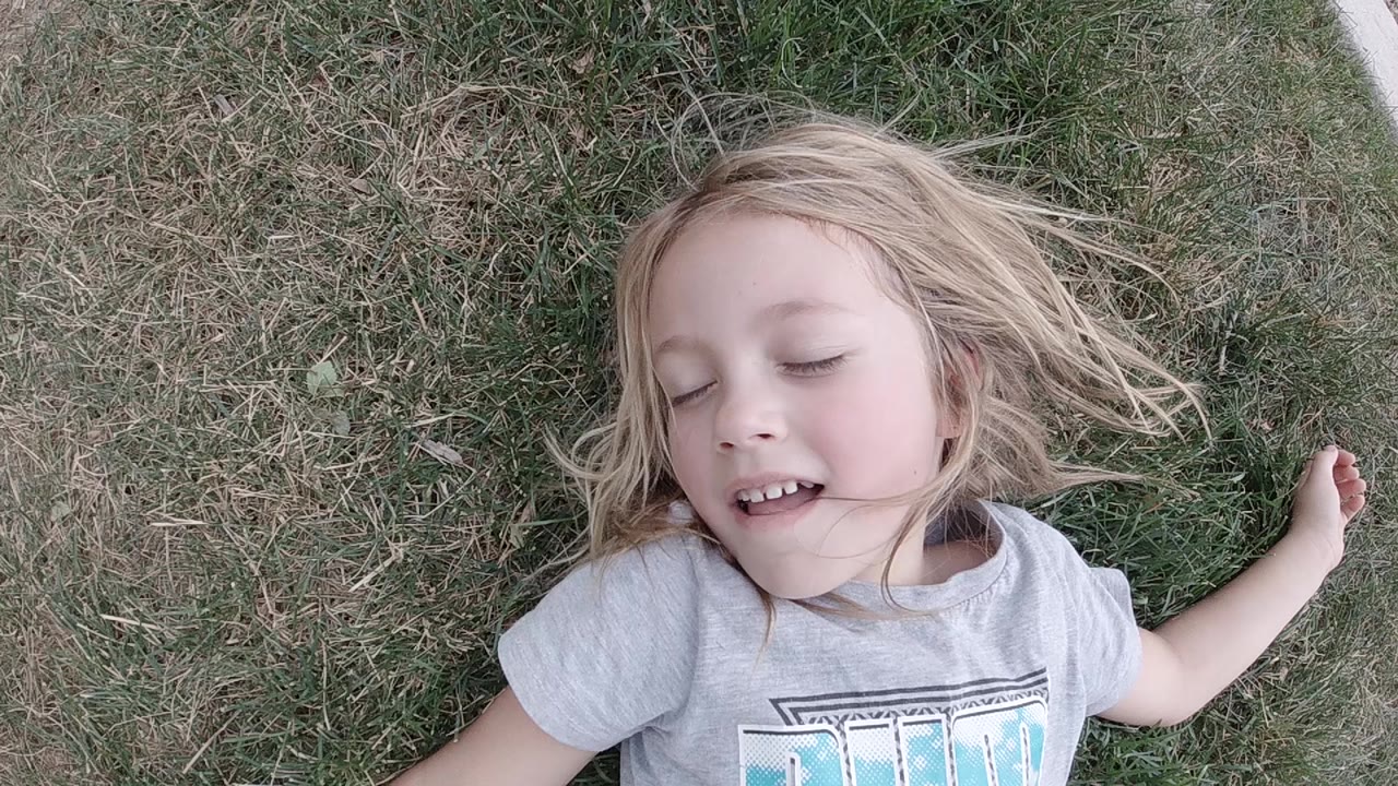 A blonde young girl lies on patchy grass in San Jose Del Cabo with her eyes closed and the top of her gray T-shirt visible.