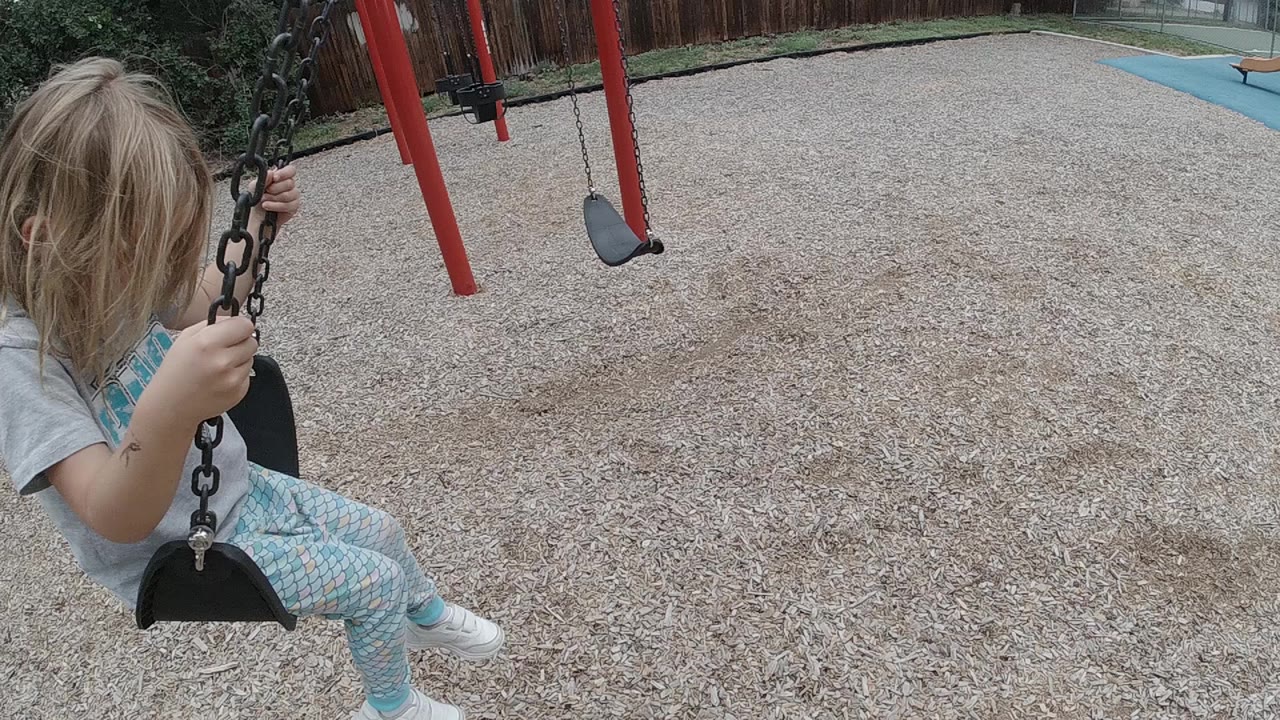 A blonde child sits close to the camera on a black chain swing in a Denver playground, with tan mulch, red swing posts, and an empty swing behind her.