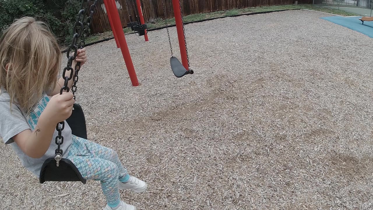 The child remains near the left edge of the frame on the swing, holding both chains as the tan playground surface and red supports fill the view.