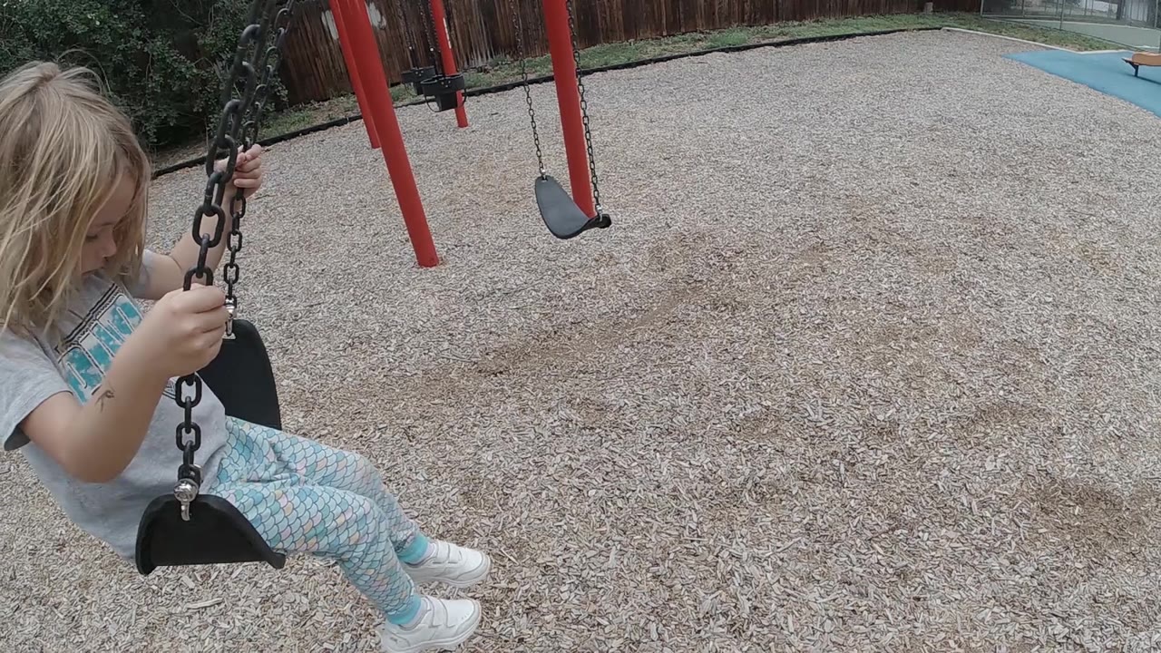 A young blonde girl sits on the black swing as it begins to move away from the camera, with red frame posts and a second empty swing in the background.