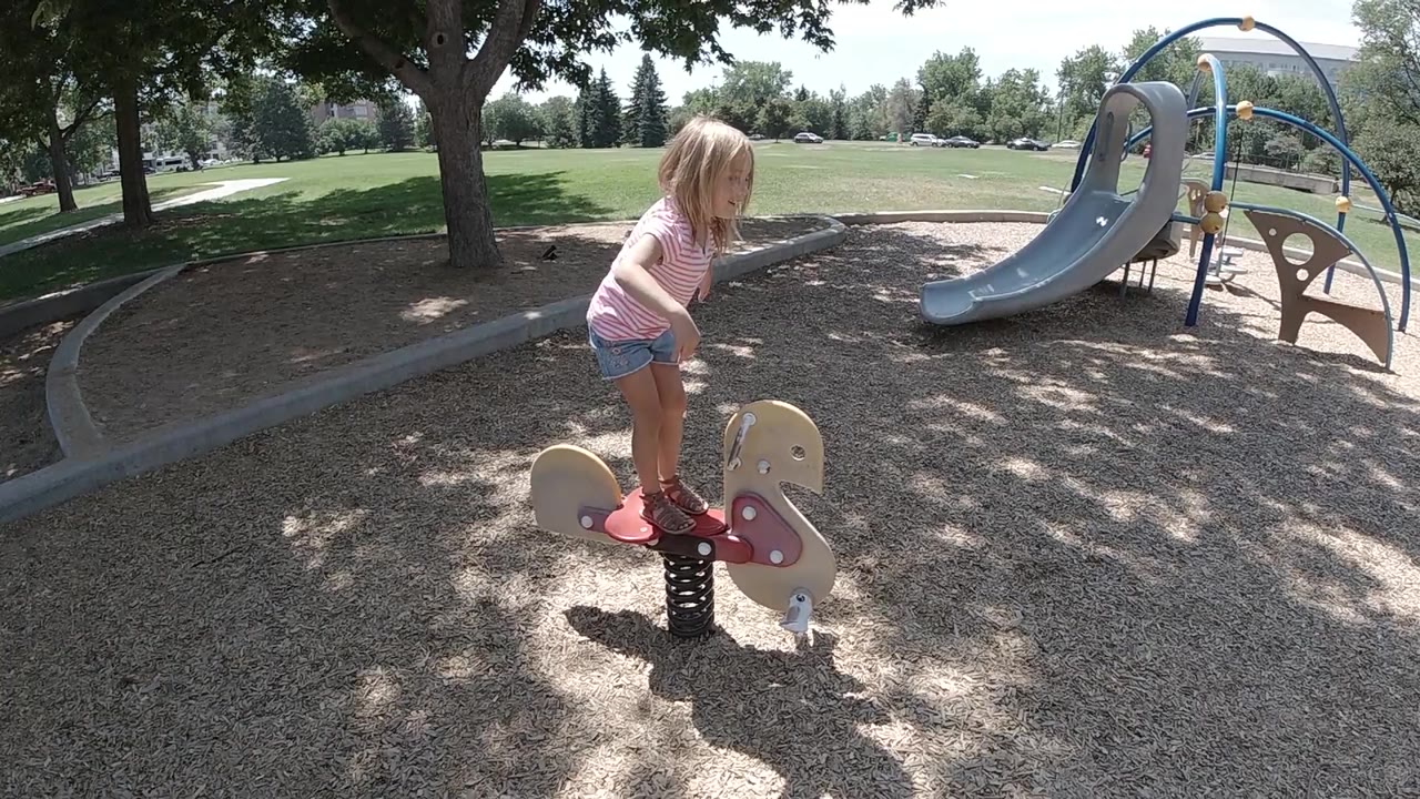 At the start, a blonde young girl stands on a red spring-mounted playground rocker over wood chips, with a blue slide and shade trees behind her.