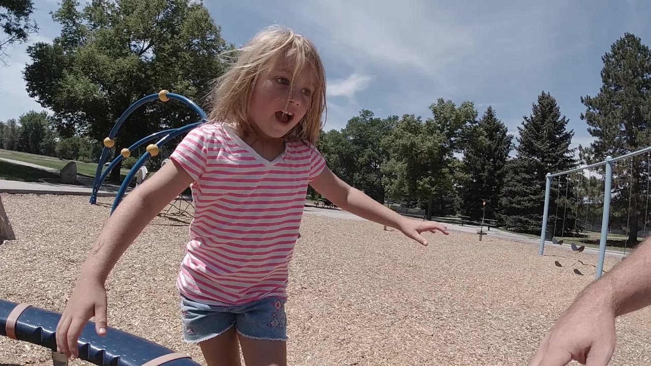 A young girl in a pink striped shirt stands close to the camera near a playground slide, with blue climbing equipment, gravel ground, trees, and sky behind her.