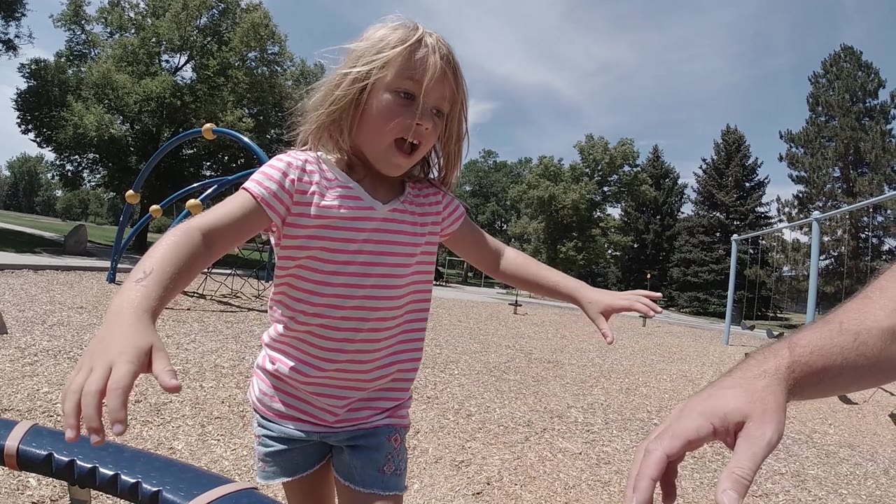The child extends both arms near the camera as an adult hand reaches in from the right, with swings and blue climbing equipment visible across the gravel playground.