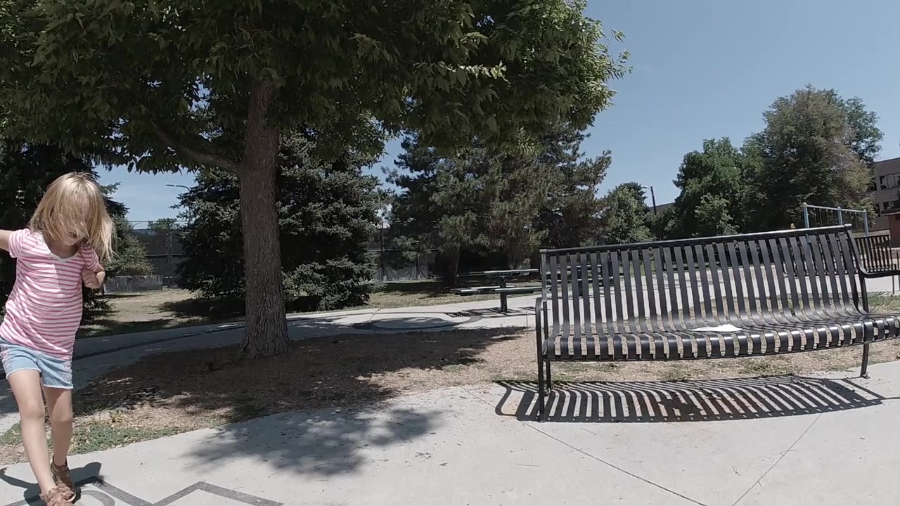 In Denver, a blonde child in a pink striped shirt steps in from the left edge beside a concrete sidewalk, metal bench, trees, and bright sky.
