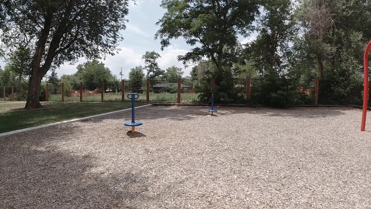 A wide Denver playground frame shows the blue spinner on gravel, a red swing support at the right edge, trees, fence, and no visible people.