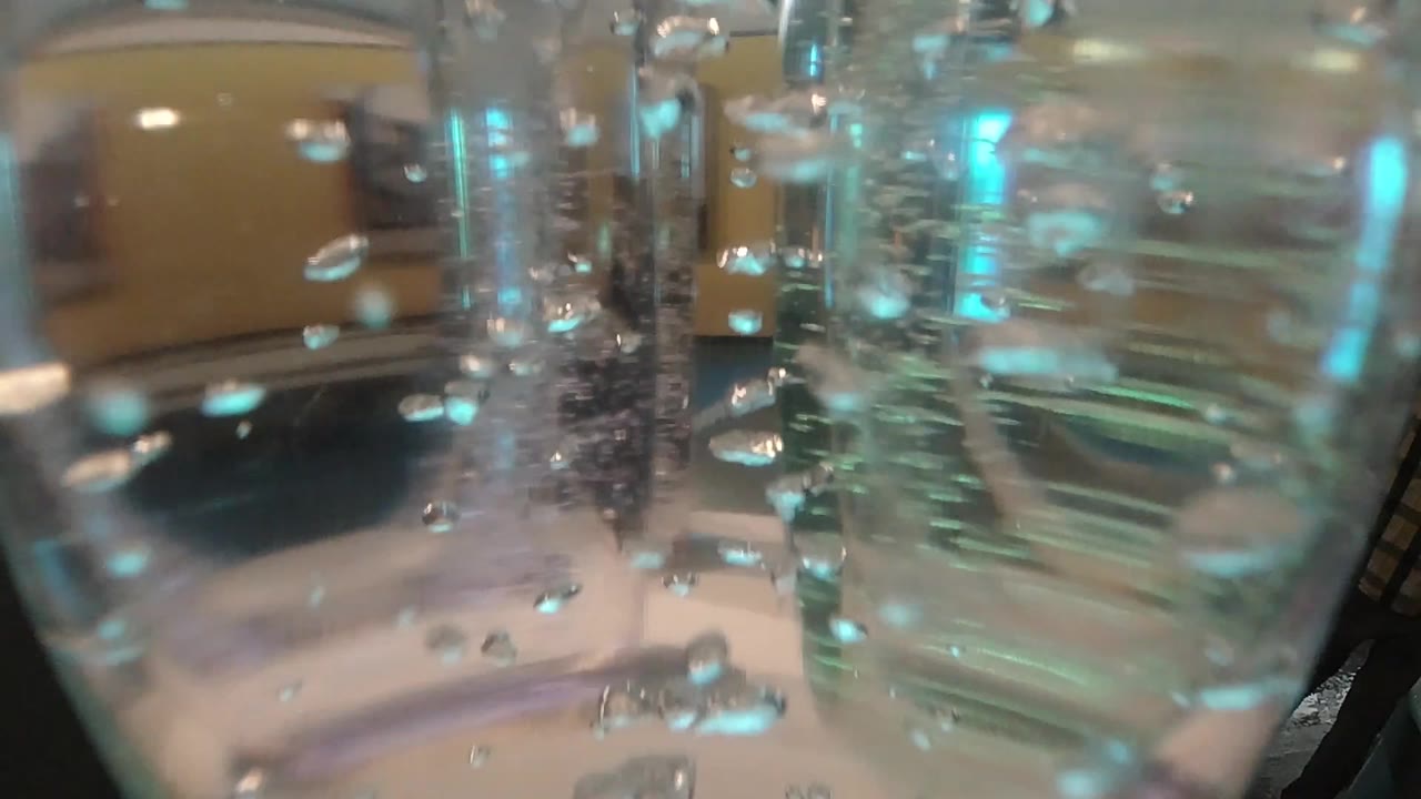 Bubbles gather across the center of a glass water container while teal light streaks reflect along the right side, with no visible people.