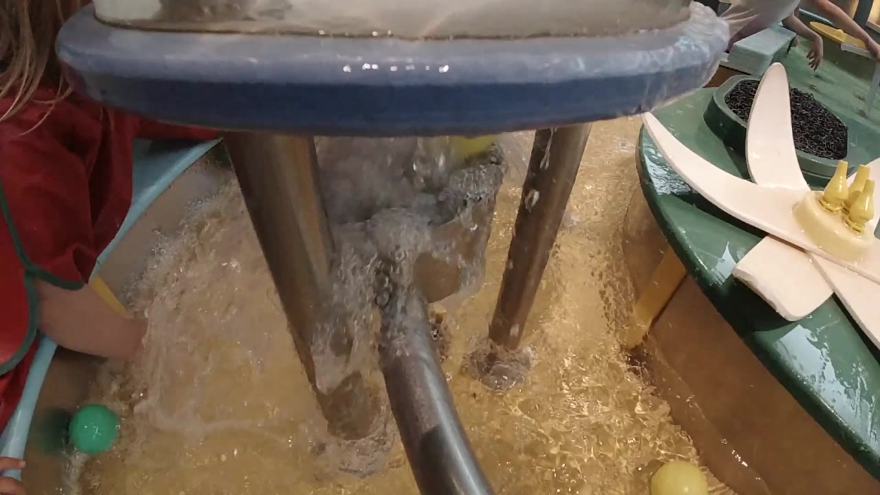 Opening frame shows foamy water pouring around vertical metal posts in a shallow basin, with part of a child in a red shirt at the left edge.